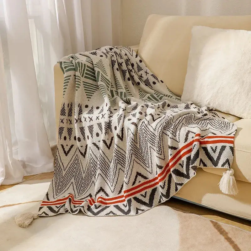 A cozy cream bohemian knitted throw blanket with bold black and green geometric patterns and orange tassels