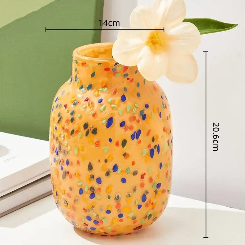 A vibrant orange DecorBites glass vase with colorful speckles and a white flower on its rim for home decor