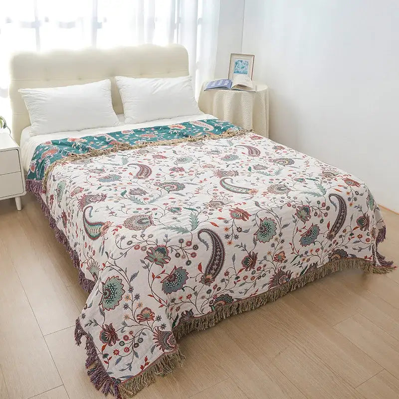 A soft cream-colored cotton bohemian throw blanket with muted paisley floral patterns and textured fringe edges