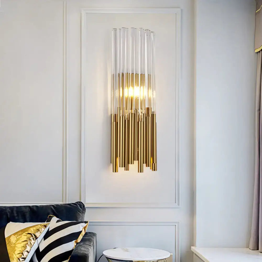 Modernist gold and clear cylinder wall light fixture with vertical glass and metal tubes emitting warm glow