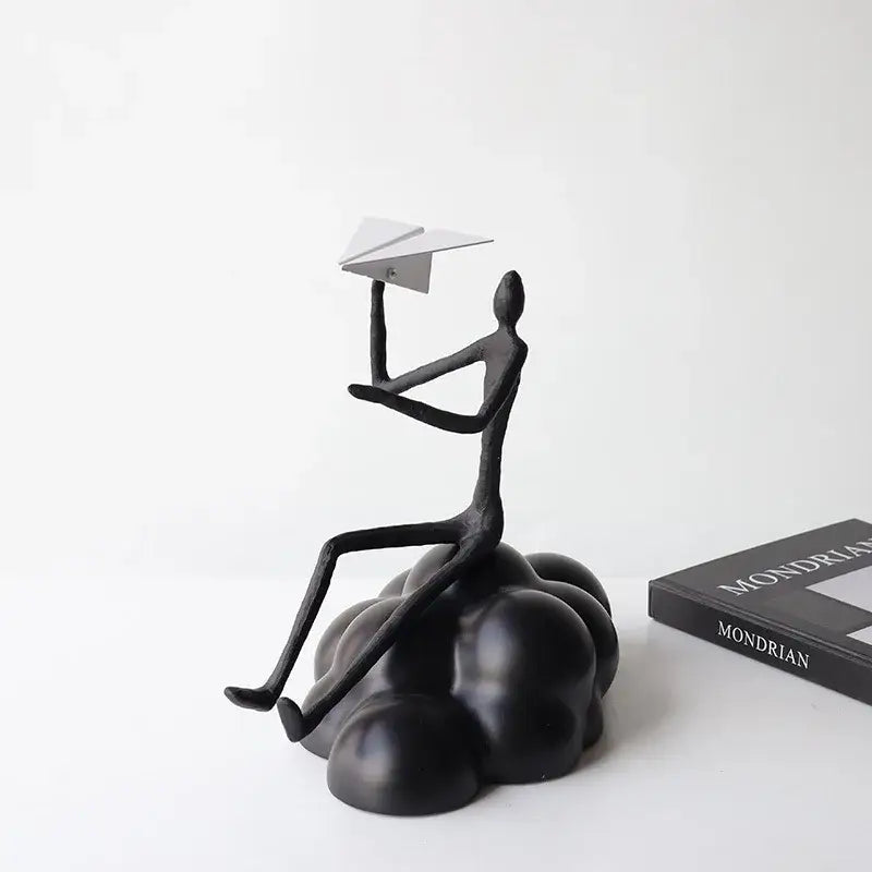 Black resin abstract figurine of a seated person holding a resin paper airplane on a paper airplane cloud base for decor
