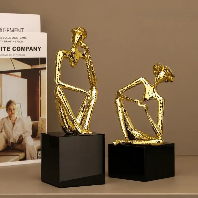 Two abstract gold hat thinker figurines seated on black square bases for living room decorbites golden hat decor