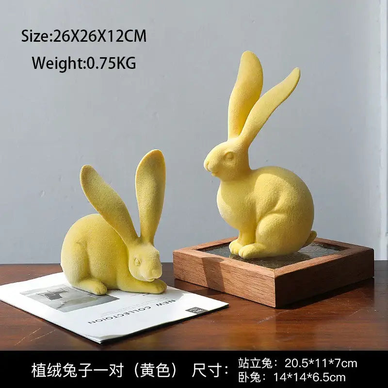 Pair of yellow plush rabbit figurines one standing one sitting beside abstract green and blue resin rabbit figurines decor