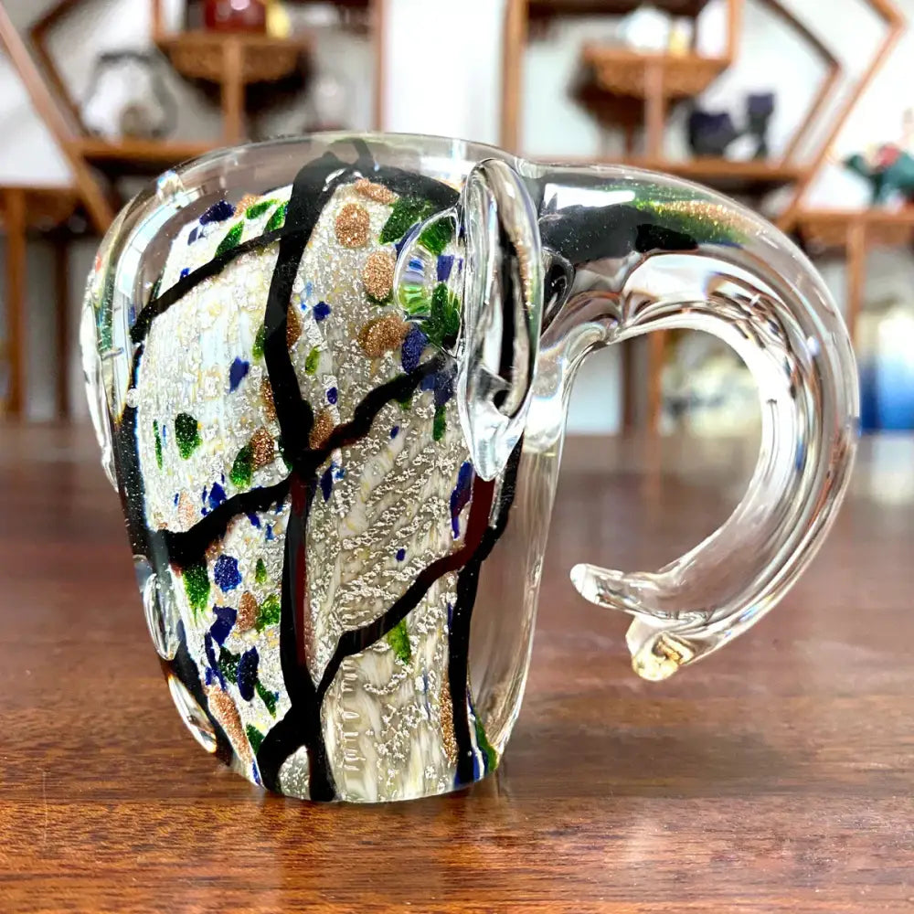 Clear glass mug with black branches green leaves and gold blue speckles next to decorbites elephant crystal glass sculpture