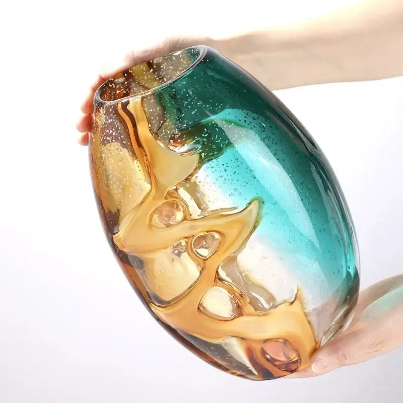 A hand holds an abstract ripple glass vase with swirling amber and teal hues and embedded bubbles for decorbites abstract ripple decor