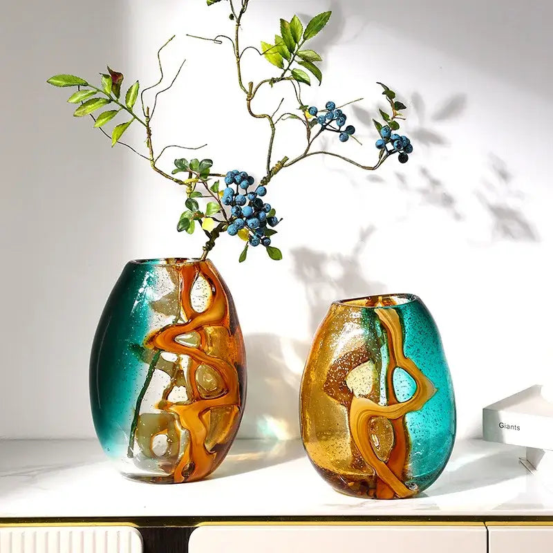 Two hand-blown abstract ripple glass vases in amber and teal with greenery and blue berries for living room decor