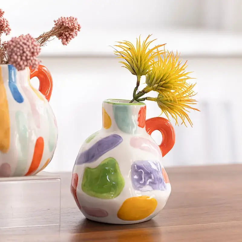 White ceramic cartoon ceramic vase teapot with colorful splatters and orange handle holding yellow spiky flower decorbites