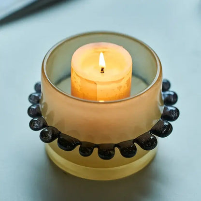A translucent amber glass cylinder vase with black beaded rim holds a lit orange candle for living room decor
