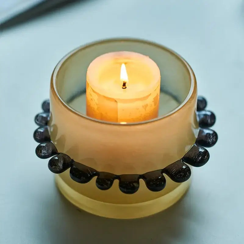 A translucent amber glass cylinder vase with black beaded rim holds a lit orange candle for living room decor