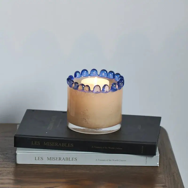A lit beige scented candle in a clear glass cylinder vase with a decorative blue beaded rim for living room decor