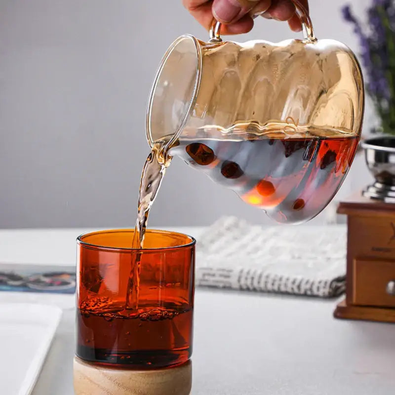A translucent amber glass coffee filter pot with textured wavy surface and gold handles pouring into orange glass