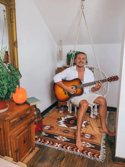 A man holding a sunburst-finish acoustic guitar in a cozy room with an american country plaid tan and blue blanket sofa cover