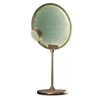 A round textured green mirror with a brass frame and stand shown with decorbites clear pink glass pendant light sphere
