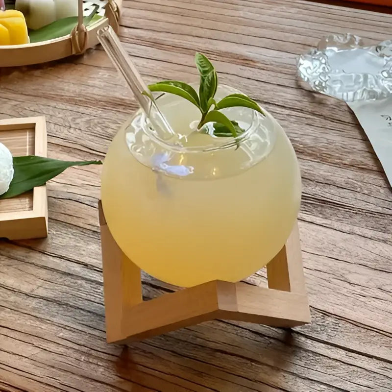 A clear glass ball shape coffee juice cocktail filled with pale yellow drink garnished with mint on a wooden stand