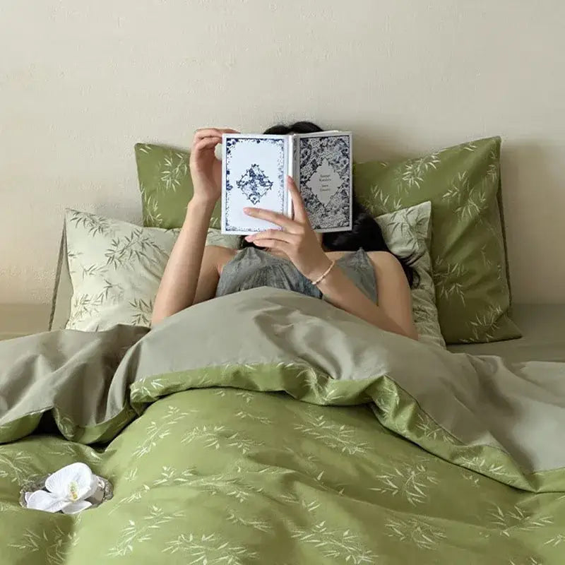 A person reclines in bed partially covered by a book on a green cotton bamboo leaf jacquard bedding set decorbites