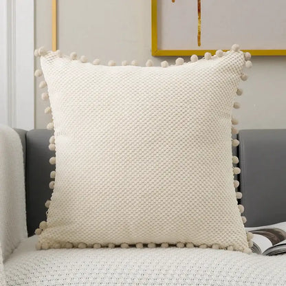 Cream-colored knit decorative pillow cover with textured woven pattern and soft pom-pom trim for living room decor