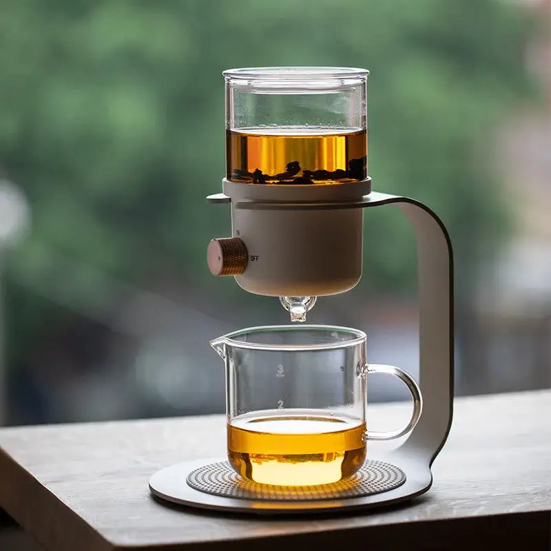 Modern glass kung fu tea pot with white ceramic base and copper knob dripping brewed tea into clear measuring pitcher