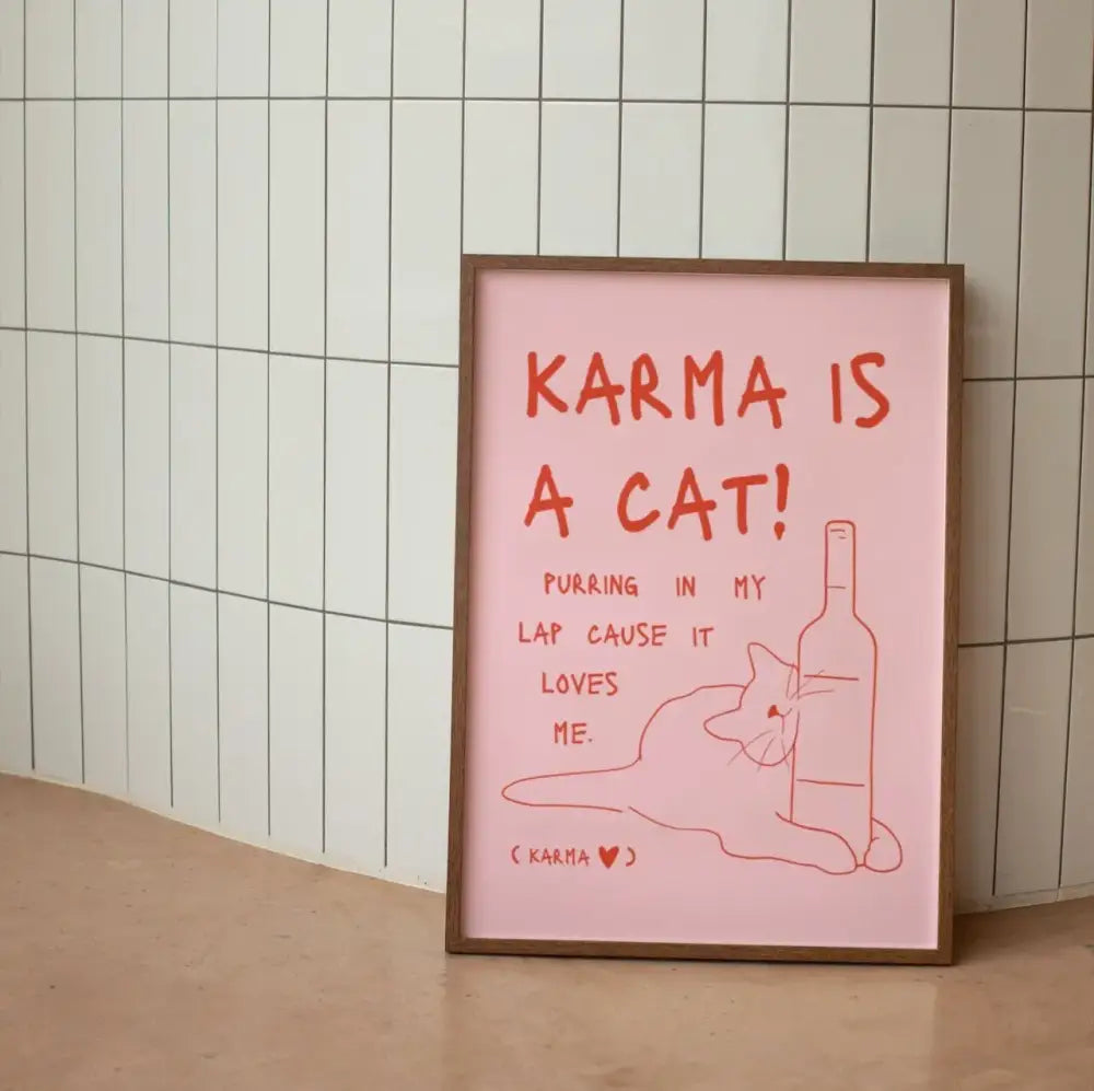 A framed pink wall art poster with red text featuring a black cat line art and the phrase karma is a cat for decor