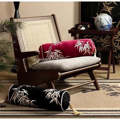 A red cylindrical bolster pillow cover with white bamboo leaves embroidery and tassel trim on a wooden chair seat