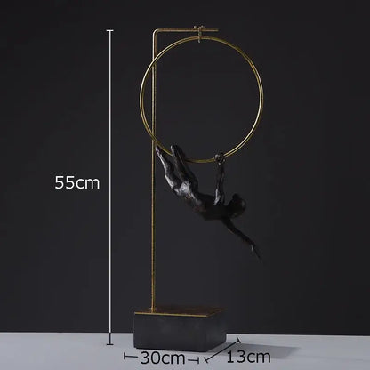 A black and gold gymnastic acrobat resin sculpture with a sleek figurine hanging from a golden hoop on a stand