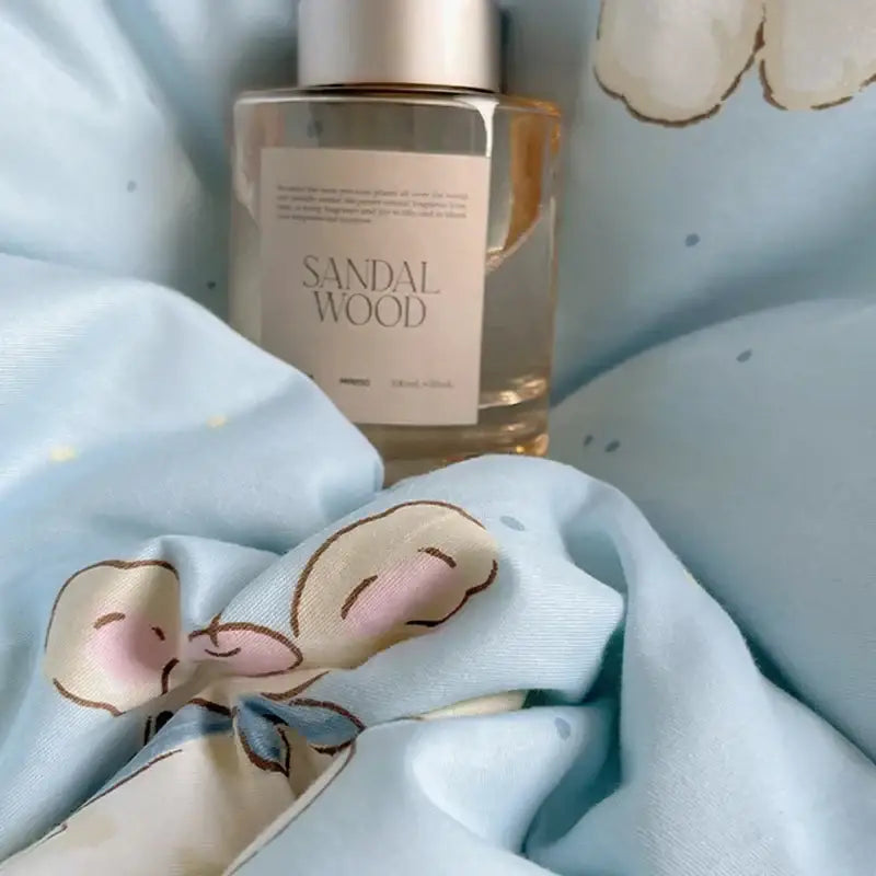 A clear glass bottle with silver cap and beige label containing amber sandalwood fragrance near blue poodle print cotton cartoon bedding set