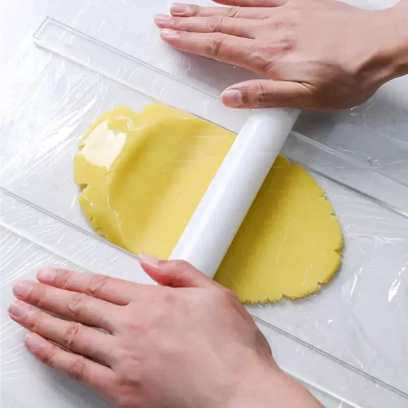 White rolling pin flattening yellow dough on clear plastic wrap with blue silicone rolling balance ruler with wood handles