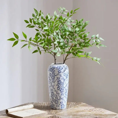A tall slender white ceramic vase with blue floral pattern holding green foliage for decorbites blue living room decor
