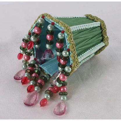 A decorative emerald green cloth jewelry box with gold beading and cascading pink red and pearl-like beads spilling out