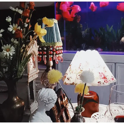 A decorative table lamp with a floral-patterned white shade and an emerald green cloth pleated lampshade with beads