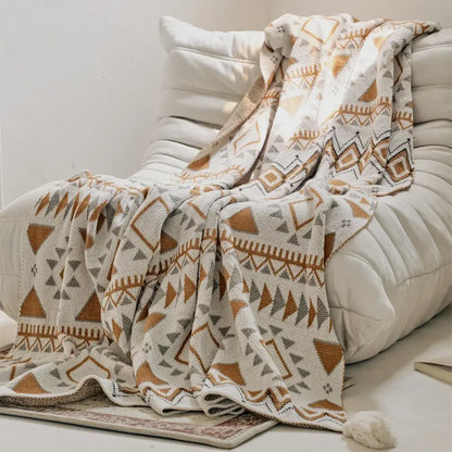 A cozy cream-colored knit throw blanket featuring a geometric pattern in warm terracotta and muted gray tones, with a soft tassel at one corner.