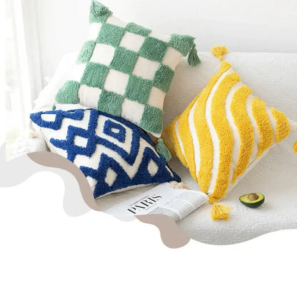 Three colorful, textured throw pillows with tassels, green and white checkered, blue geometric pattern on cream base, and yellow and white striped, all made of plush yarn.