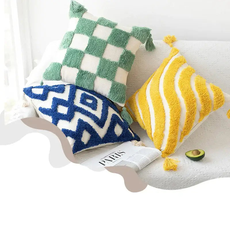 Three colorful, textured throw pillows with tassels, green and white checkered, blue geometric pattern on cream base, and yellow and white striped, all made of plush yarn.