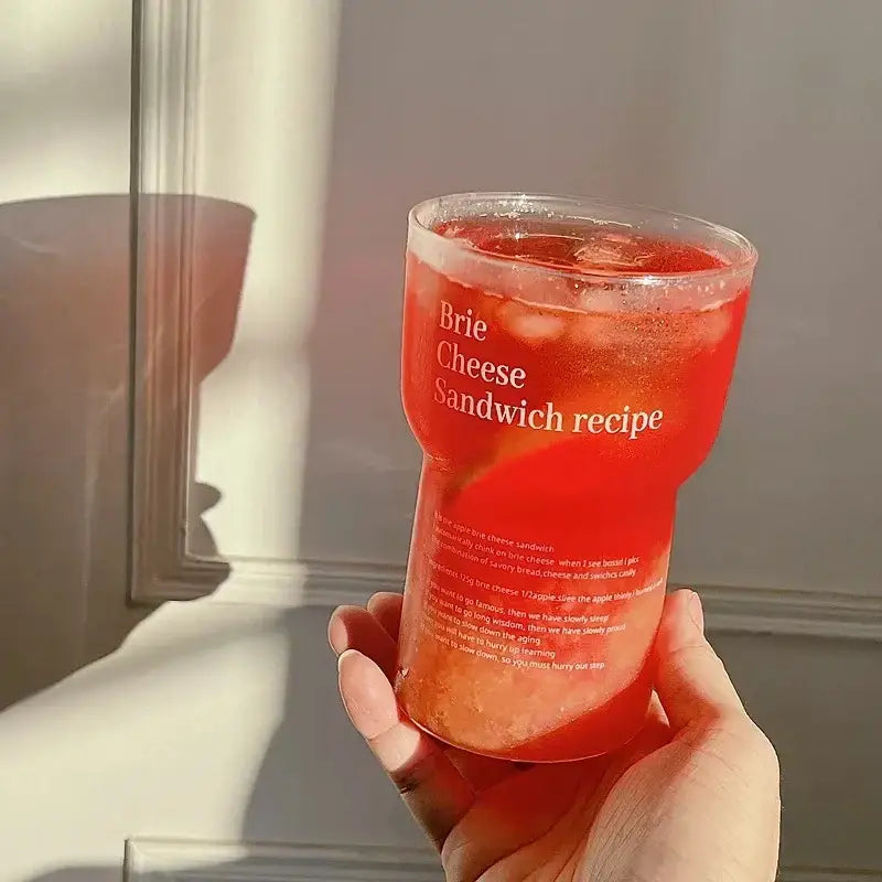Translucent red plastic cup with brie cheese sandwich recipe in white text filled with ice and reddish beverage used as glass coffee mug