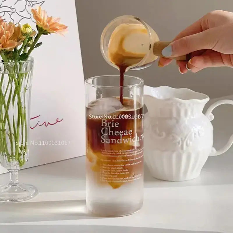 A clear glass coffee mug with frosted base and brie cheesecake sandwich printed text in black font with straw