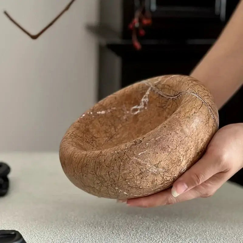 A hand holds a rustic brown and gray nest marble bowl ashtray with a cracked textured surface and subtle metallic glints