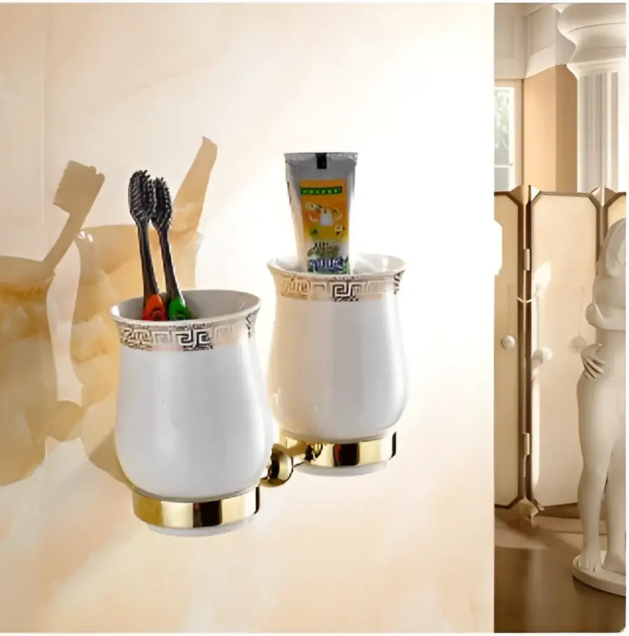 White ceramic toothbrush holder with gold trim and greek key pattern holding black toothbrushes in bathroom hardware set