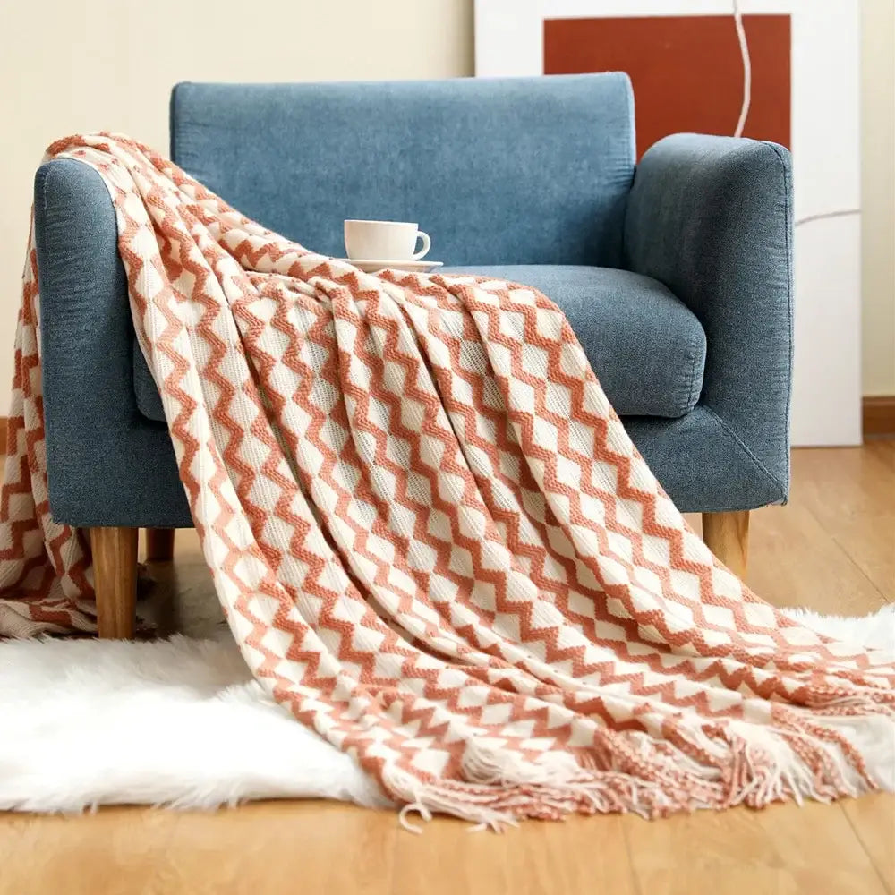 A cozy knitted sofa blanket in peach and cream zigzag pattern with fringed edges draped over a blue armchair