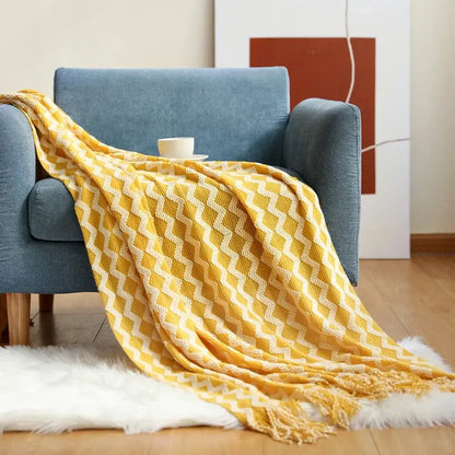 A cozy knitted sofa blanket in mustard yellow with white zigzag pattern and fringed edges draped on a blue armchair