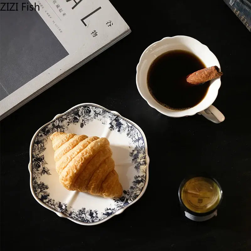 A white ceramic plate with black floral edges holds a croissant beside a grey blue rose ceramic cup and saucer set