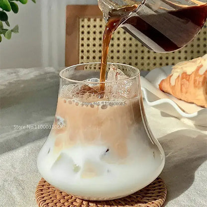 A clear glass cold brew latte drinkware set with a wooden coaster features a pitcher pouring dark coffee into iced milk