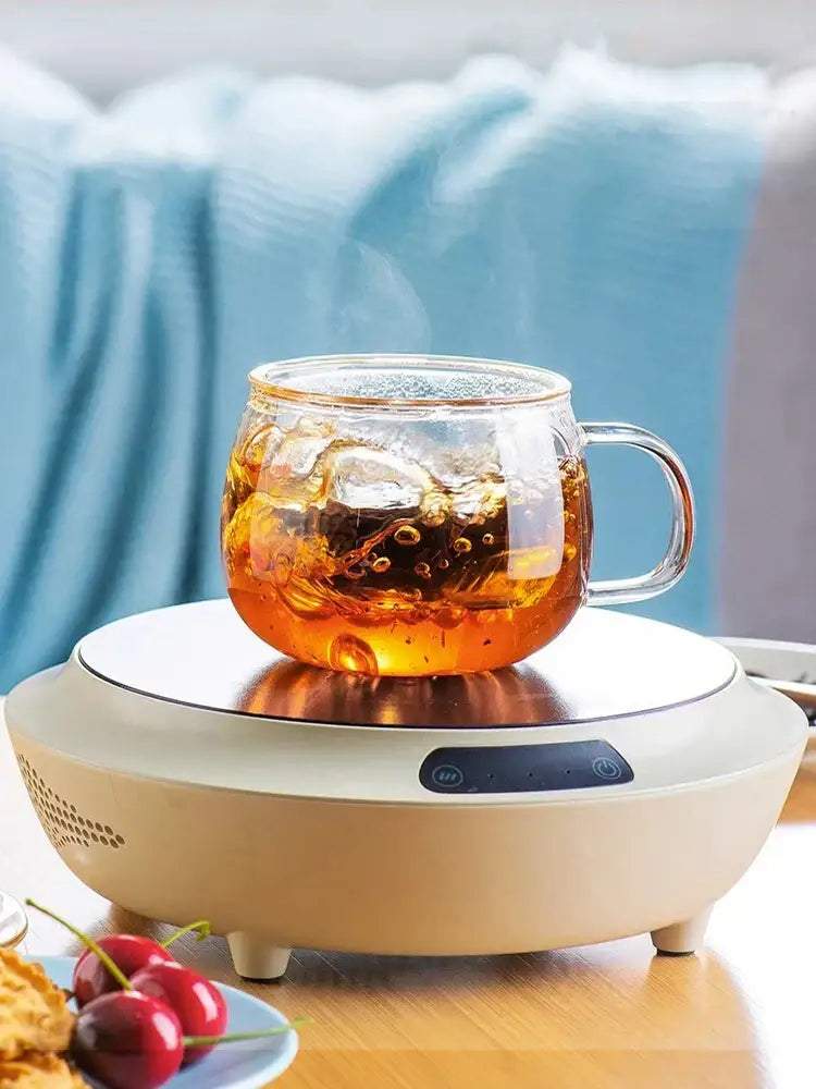 Clear glass floral tea cup with handle filled with amber tea and ice cubes on off-white electric heating base