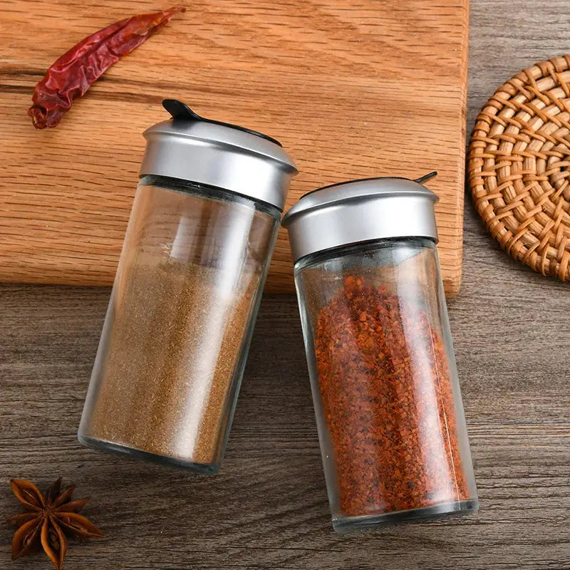 Two clear glass rotating spice jars with silver lids containing brown seasoning and red chili powder for kitchen use