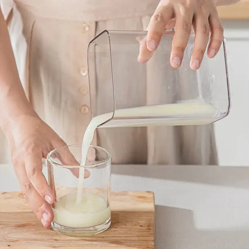 Clear rectangular glass pitcher pouring milk into glass next to clear greek yogurt maker with white lid and mesh filter