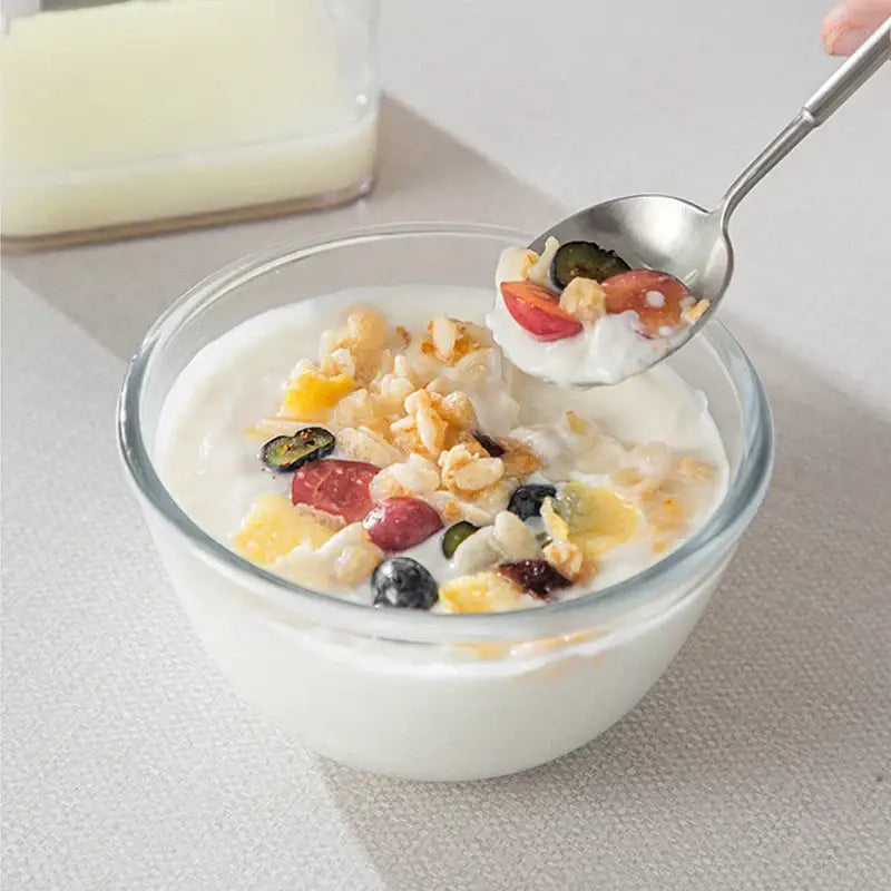 A clear glass bowl with creamy greek yogurt topped with mixed fruit and granola beside a clear greek yogurt maker with white lid and mesh filter