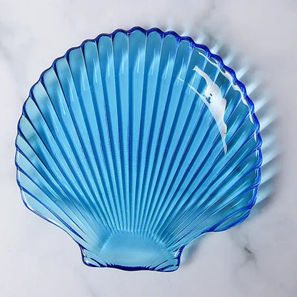 Translucent blue glass dish plate shaped like a scallop shell with ridged grooves and a glossy finish for dining use
