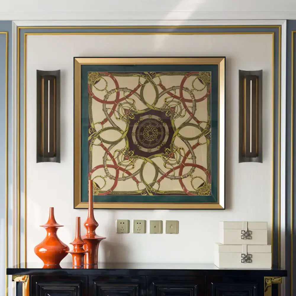 A framed hermès scarf print with red gold and beige tones displayed beside a contemporary black led wall lamp with metal shade
