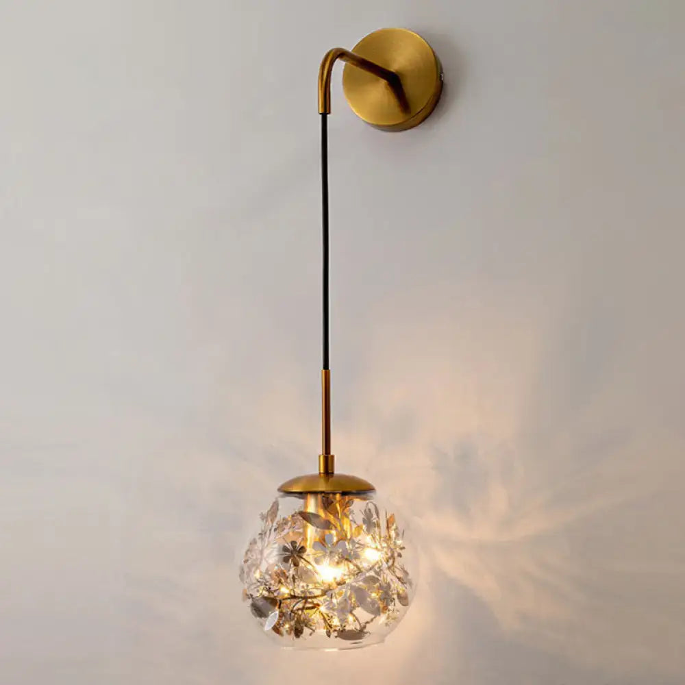 Contemporary brass wall lamp with clear glass globe and foil flower decor suspended by a black cord for bedroom use
