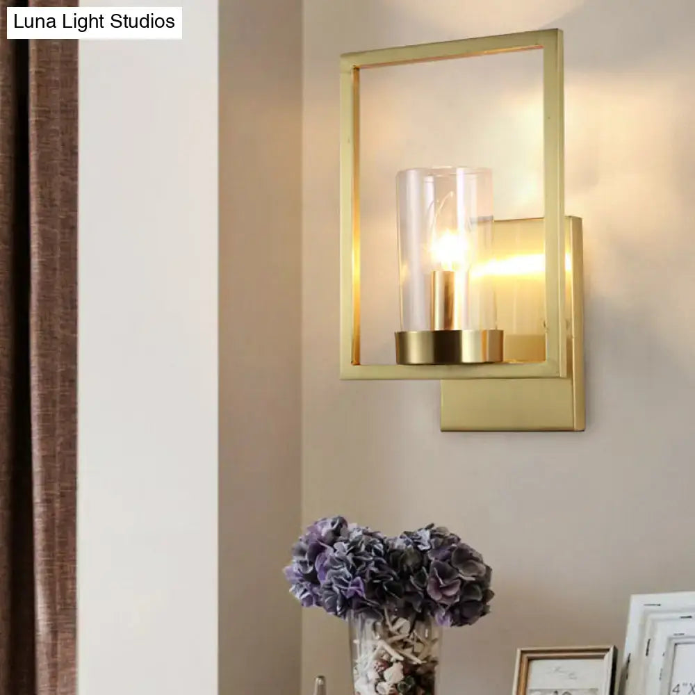 Gold-framed contemporary brass wall light with clear glass cylinder shade and illuminated bulb for bedroom lighting fixture