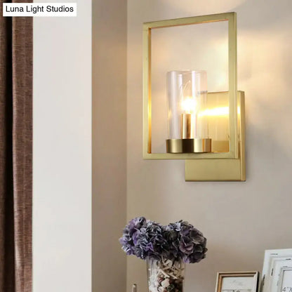 Gold-framed contemporary brass wall light with clear glass cylinder shade and illuminated bulb for bedroom lighting fixture