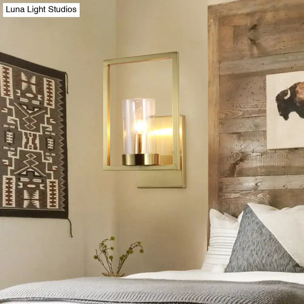 Modern brass-framed wall light with clear glass cylinder housing an exposed bulb for contemporary bedroom use